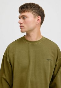 Olive zielony, oversize'owy sweter z żeberkowym dekoltem i haftowanym napisem "Codzienna rutyna" w kursywie na piersi. Miękka faktura materiału.