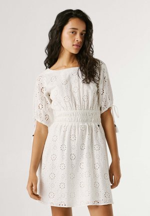 Donna con lunghi capelli ricci scuri che indossa un vestito bianco in sangallo con maniche a sbuffo e vita stretta, in piedi contro uno sfondo chiaro uniforme.