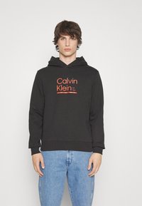 Black hoodie made of cotton with a kangaroo pocket. Features a red Calvin Klein logo and established date, ribbed cuffs, and hem.
