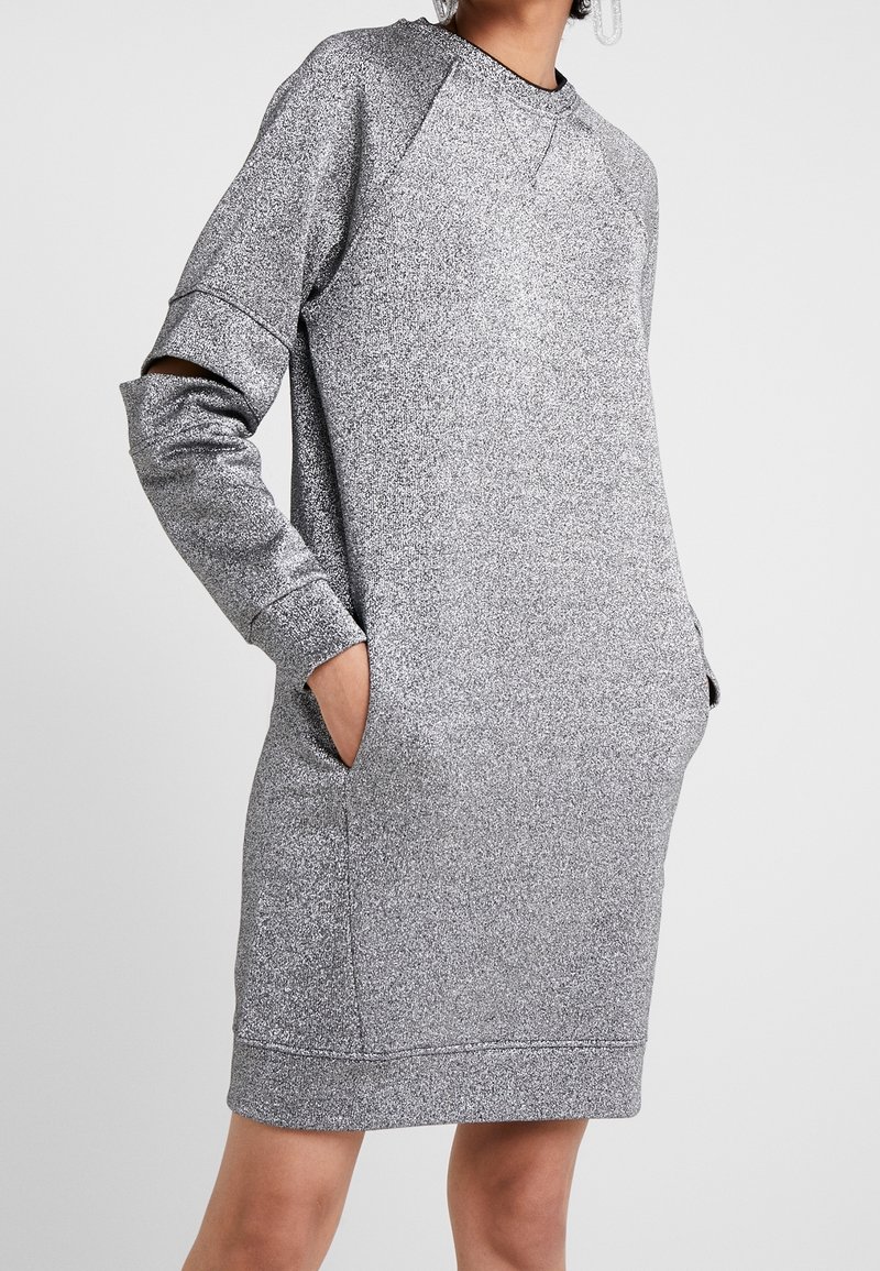Personne portant une robe longue argentée scintillante avec un détail découpé sur une manche et les mains dans les poches latérales.