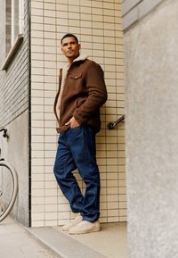 Chaqueta de mezcla de lana marrón con cuello de borreguito, combinada con pantalones de mezclilla azules y zapatillas beige claro, posicionada contra una pared de azulejos.