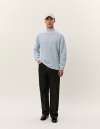 Pullover lavorato a maglia azzurro chiaro con collo alto, abbinato a jeans dritti scuri e scarpe nere, su uno sfondo neutro.