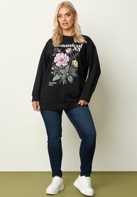 Svart sweatshirt med blommigt grafiskt mönster i rosa och gult, kombinerad med mörkblå jeans och vita sneakers. Avslappnad passform och lång ärm.