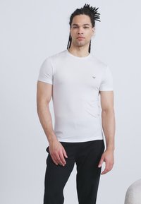 Άνδρας με dreadlocks που φοράει εφαρμοστό λευκό κοντομάνικο μπλουζάκι και μαύρο παντελόνι, στέκεται μπροστά σε απλό φωτεινό φόντο.