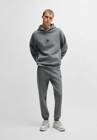 Grijze hoodie met reliëf logo, gecombineerd met bijpassende grijze joggers. Beide gemaakt van een gladde stof. Witte sneakers maken de outfit compleet.