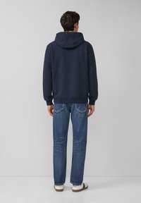 Granatowa teksturowana bluza z kapturem, zestawiona z prostymi jeansami i casualowymi sneakersami, widziana od tyłu.