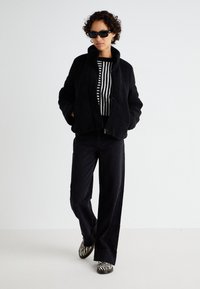 Jachetă de fleece neagră cu guler înalt, bluză cu mânecă lungă în dungi negre și albe, pantaloni negri cu croială largă și pantofi cu un model de zebre în alb și negru.