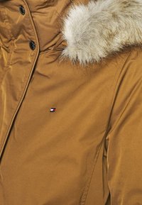 Bruine winterjas met een met bont gevoerde capuchon, zwarte drukknoopknopen en een klein rechthoekig rood, wit en blauw logo op de borst.