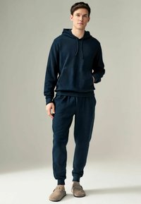Navyblauwe katoenen hoodie met koordjes en een voorzak, gecombineerd met bijpassende broek met elastische tailleband en taps toelopende boorden. Lichtgrijze schoenen.