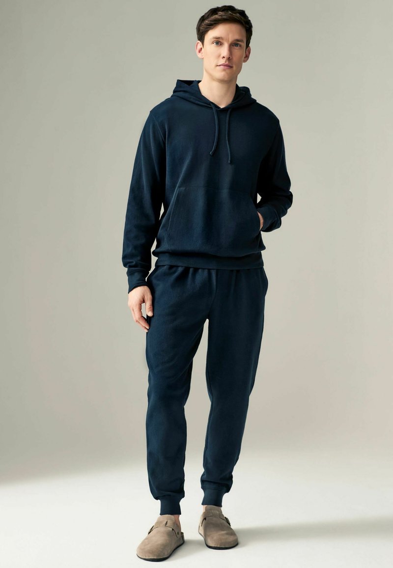 Navyblauwe katoenen hoodie met koordjes en een voorzak, gecombineerd met bijpassende broek met elastische tailleband en taps toelopende boorden. Lichtgrijze schoenen.