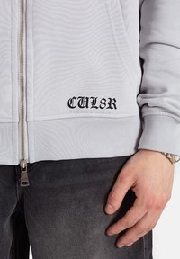 Jasnoszara bluza z kapturem na zamek błyskawiczny wykonana z teksturowanej tkaniny, z dużym czarnym nadrukiem "CUL8R" po lewej stronie oraz srebrnym zamkiem.