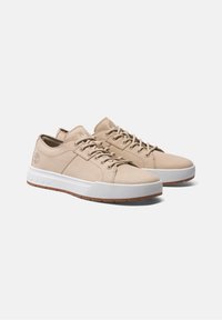 Beige lädersneakers med en vit gummisul, platta snören och en logodetalj på sidan. Har en slät yta och rundad design.
