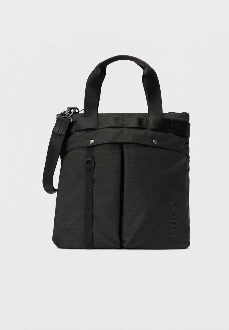 Svart stoff totebag med doble håndtak, frontlommer, avtakbar skulderreim og preget "HUGO"-logo nederst til høyre.
