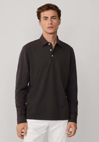 Polo a maniche lunghe marrone scuro con colletto classico, placca con tre bottoni e una texture morbida. Indossato con pantaloni di colore chiaro.