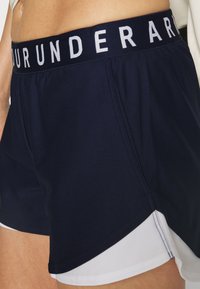 Navy siniset urheilushortsit, joissa on valkoinen alaosa. Täydentää voimakas musta vyötärönauha, jossa lukee "UNDER ARMOUR". Valmistettu kevyestä kankaasta.