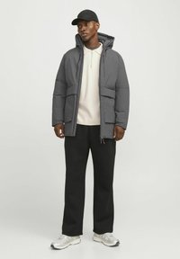 Grå pufferjakke med hætte, to forlommer og lynlåslukning, båret over en lys sweater og sorte joggers, parret med grå sneakers.