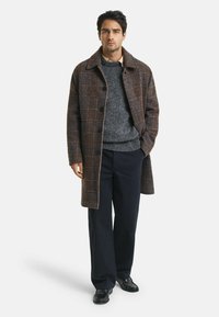 Mann trägt einen braunen karierten Mantel, einen grauen Pullover, eine marineblaue Hose und schwarze Schuhe und steht mit einer Hand in der Tasche vor einem weißen Hintergrund.