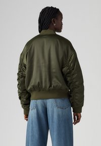 Olive green satin bomber jacket with ribbed cuffs and hem, featuring gathered sleeves and a relaxed fit, paired with light blue wide-leg jeans.