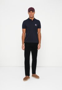 Marineblå polo skjorte med en liten logo, kombinert med svarte bukser, brune slip-on sko og en burgunder caps, står mot en ensfarget bakgrunn.