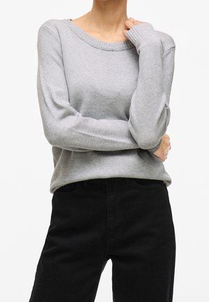 Person wearing light grey long-sleeve knit sweater with ribbed neckline and black pants, arms crossed, with one hand touching their neck.