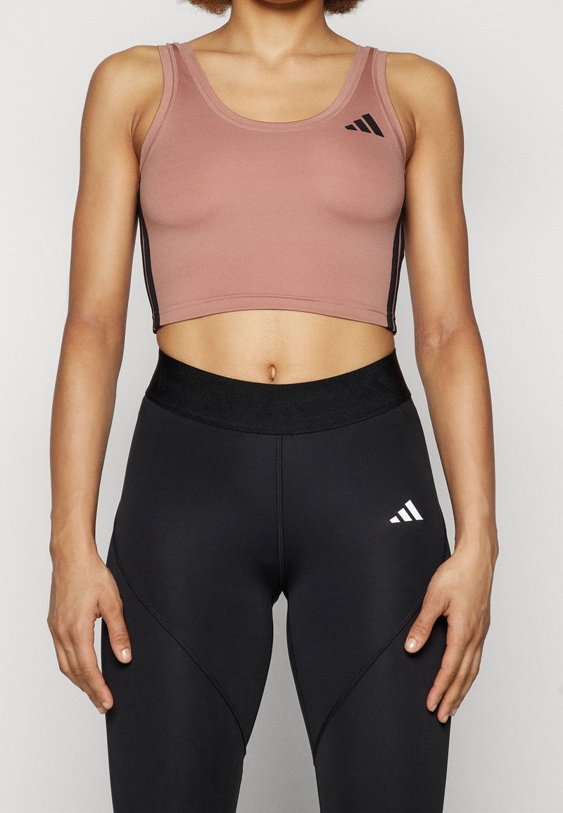 Femme portant un top de sport court mauve et des leggings noirs taille haute avec des logos blancs et noirs sur un fond uni.