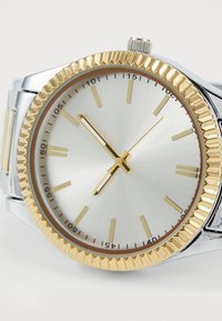 Montre argentée avec une lunette en or texturée, un cadran argenté, des index horaires dorés et une couronne de remontoir. Possède un bracelet en maillons.
