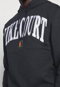 Sudadera negra con letras blancas "NIKE COURT", un pequeño logo de Nike multicolor y un bolsillo tipo canguro. Fabricada en una suave mezcla de algodón.