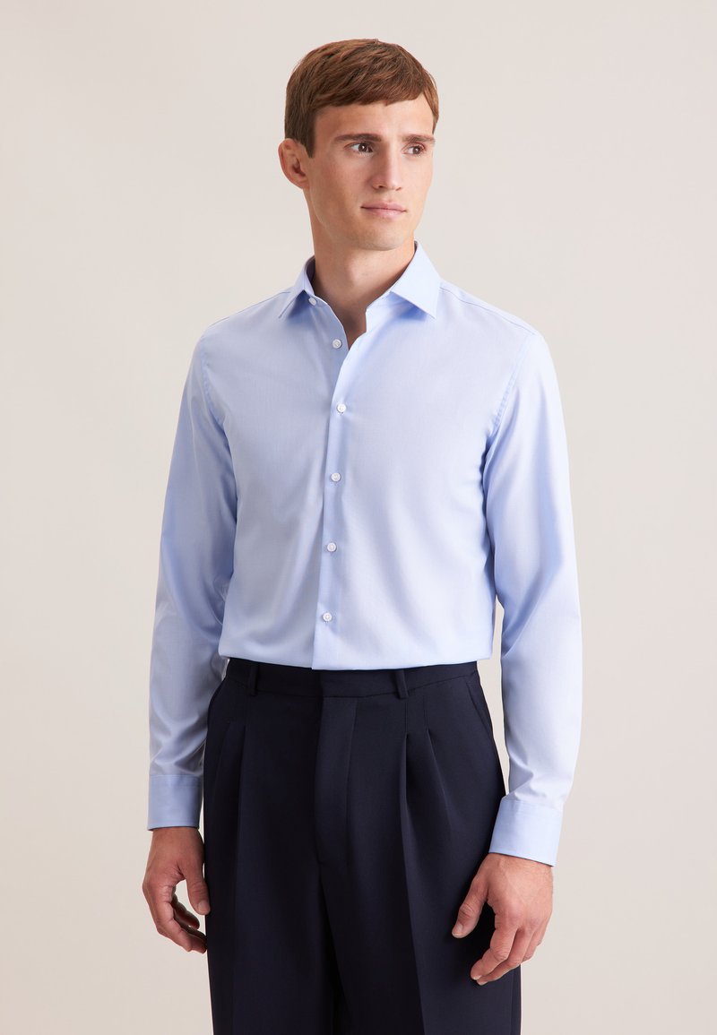 Homme portant une chemise bleue claire à manches longues boutonnée et un pantalon foncé, debout devant un fond beige uni, regardant sur le côté.