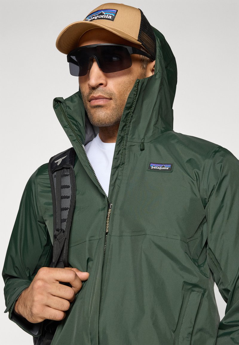 Veste vert foncé avec un col haut, une fermeture éclair et le logo Patagonia. Les bretelles d'un sac à dos noir sont visibles. Casquette marron avec un dos en mesh et un logo. Lunettes de soleil foncées.