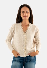Femme aux longs cheveux noirs portant un cardigan beige à boutons avec des détails en dentelle et un jean bleu, les mains dans les poches, fond neutre.