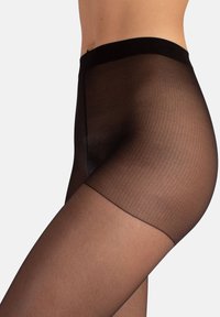Primer plano de una persona que lleva pantys negros transparentes con una cinturilla reforzada más oscura y la parte superior del muslo, contra un fondo liso.