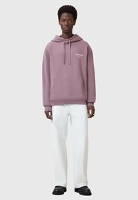 Felpa lavanda oversize con tasca a marsupio, cappuccio con lacci e logo bianco. Abbinata a pantaloni bianchi a gamba larga e scarpe nere.