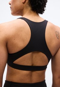 Άτομο που φοράει μαύρο αθλητικό σουτιέν racerback, δείχνοντας την πλάτη και τον ώμο με ένα μικρό τατουάζ στην δεξιά ωμοπλάτη.