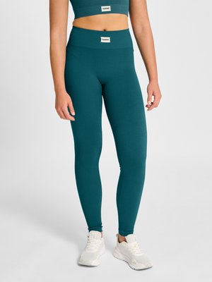 Vrouw draagt teal geribbelde leggings met hoge taille en bijpassend cropped topje, staand met witte sneakers tegen een effen achtergrond.