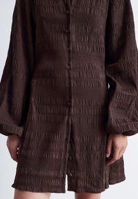 Braunes Kleid aus strukturiertem Stoff mit vertikalen Streifen. Verfügt über eine Knopfleiste und lockere, lange Ärmel.