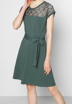 Femme portant une robe vert foncé jusqu'aux genoux avec un empiècement en dentelle, des manches courtes et une ceinture en tissu nouée en nœud à la taille.