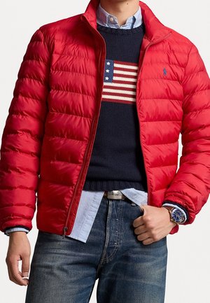 Homme portant une veste matelassée rouge sur un pull bleu marine avec un motif du drapeau américain, une chemise bleu clair, un jean bleu et une montre-bracelet.