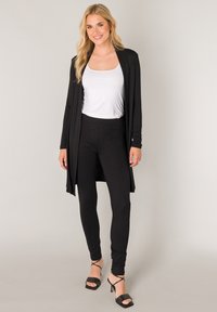 Zwarte lange cardigan over een wit topje, gecombineerd met zwarte legging en zwarte sandalen met bandjes. Zachte textuur, eenvoudig ontwerp, geen zichtbare patronen.