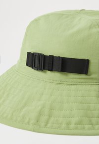 Chapeau de seau vert clair en tissu texturé, doté d'une sangle noire ajustable et de détails de couture visibles le long de la bordure.
