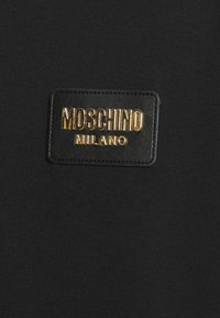 Black knit fabric with a rectangular black leather patch featuring embossed gold lettering "MOSCHINO MILANO." Smooth texture, minimalistic design.