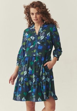 Ung kvinde med krøllet hår iført en mørkeblå blomsterkjole med grønne og blå blade, med hånden i lommen, stående foran en ensfarvet baggrund.