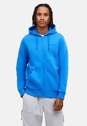 Jonge man met gevlochten haar, gekleed in een felblauwe hoodie met rits en lichtgrijze cargobroek, staand met handen in de zakken.