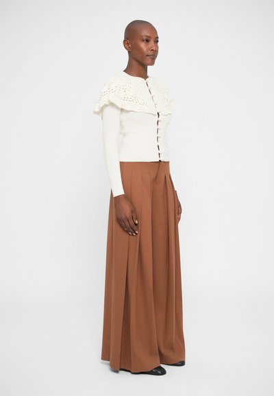 Off-white pletený kardigán s čipkovým golierom, v kombinácii s vysokým pásom a širokými hnedými nohavicami. Hladká textúra, zapínanie na gombíky, elegantný dizajn.