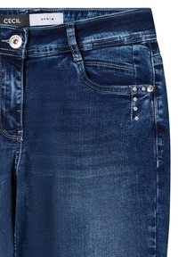 Pantalones vaqueros de mezclilla azul oscuro con textura suave, que presentan un bolsillo delantero y tachuelas decorativas plateadas en el borde del bolsillo. Etiqueta de la marca CECIL visible.