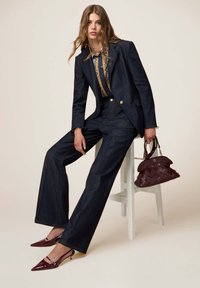 Traje de denim con un blazer estructurado y pantalones de pierna ancha en azul oscuro. Tacones de charol burdeos tipo slingback y un bolso tejido burdeos.