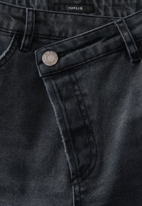 Donkerdenim jeans met een lichte textuur, voorzien van een zilveren knop en een gestikte OPUS-label op de tailleband. Close-up van de details.