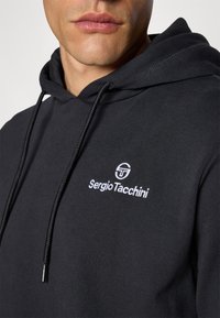 Svart hettegenser med snorer, med en hvit brodert logo "Sergio Tacchini" på brystet. Mykt bomullsmateriale.