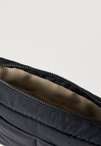 Μαύρο καπιτονέ θήκη με ανοιχτό φερμουάρ που αποκαλύπτει εσωτερική επένδυση σε μπεζ χρώμα, πάνω σε απλό ανοιχτόχρωμο φόντο.