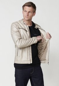 Bež jakna iz umetnega usnja s prešito notranjostjo, žepom na zip in stoječim ovratnikom. Model nosi črno majico pod jakno.