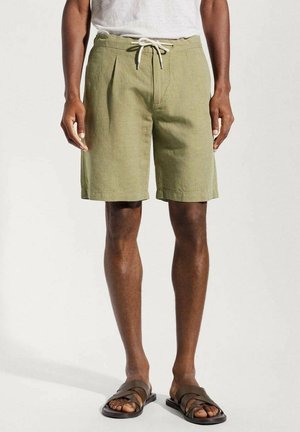 Shorts en lin vert clair avec des plis, taille à cordon, et poches latérales, associés à des sandales marron à lanières, portés par une personne debout.